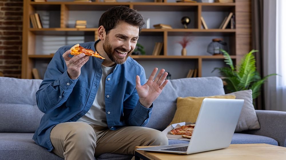 Happy adult male eating a piece of pizza while having a video chat on his laptop in a cozy living room setting.